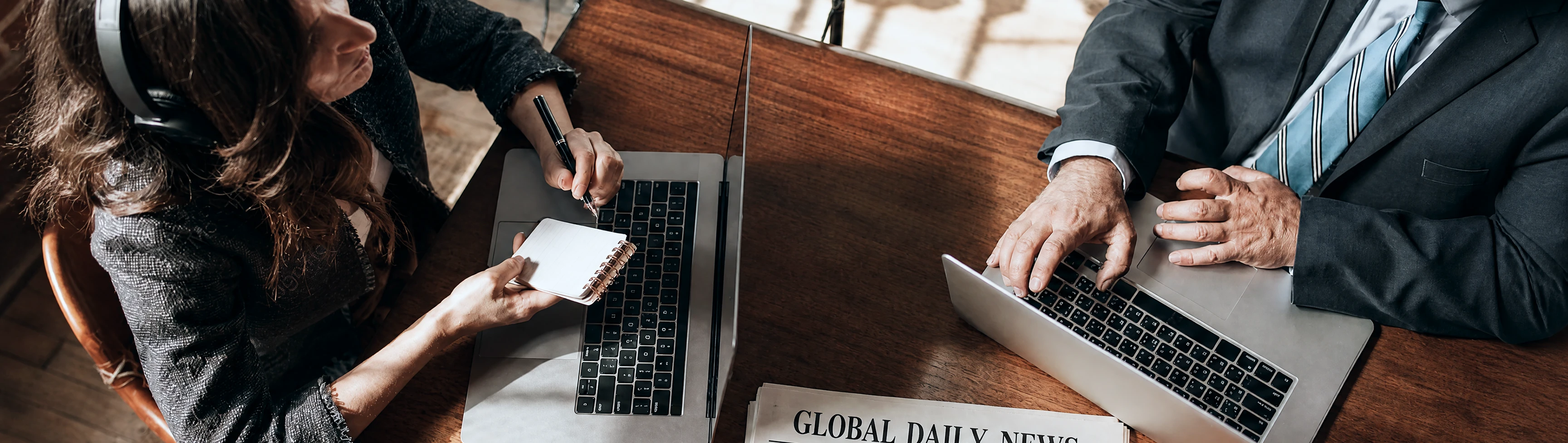 Two professionals collaborating at a desk with laptops and a newspaper.